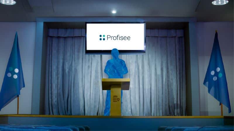 A person stands at a podium in a room resembling a house of parliament with flags bearing the Profisee logo stand on either side of them.